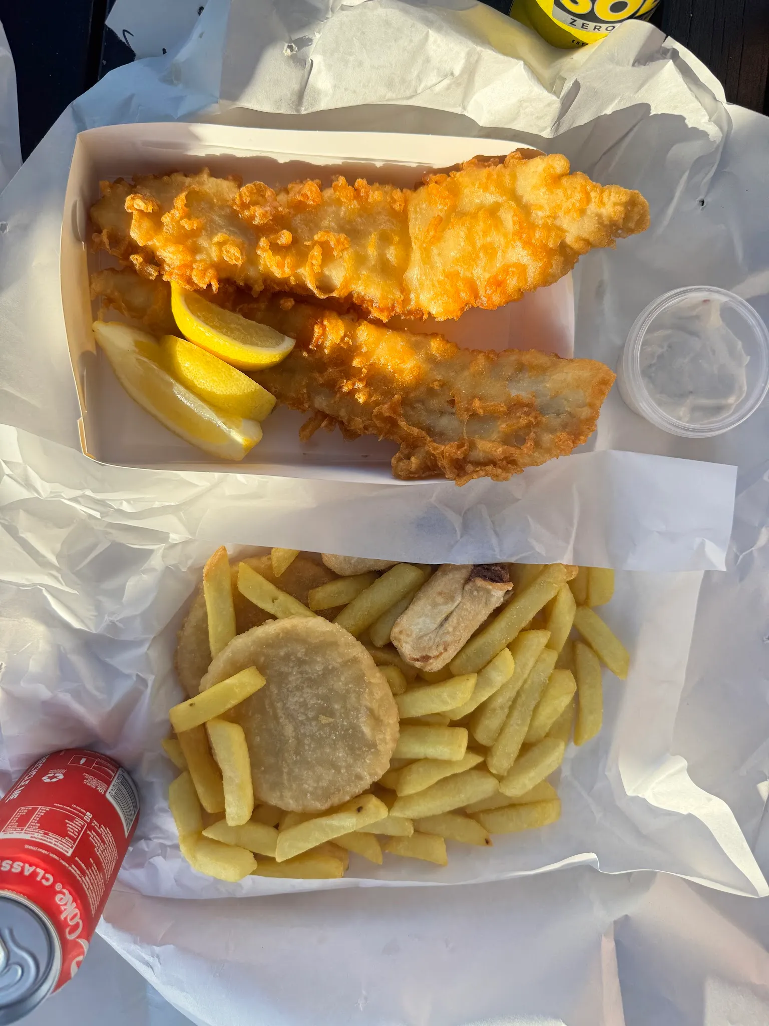 Two takeaway boxes of battered fish with lemon, chips and a potato cake